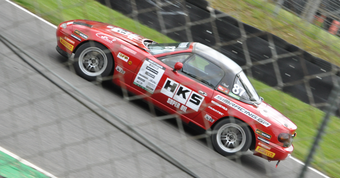 Red number 87 Mazda MX-5 on track behind a safety fence.