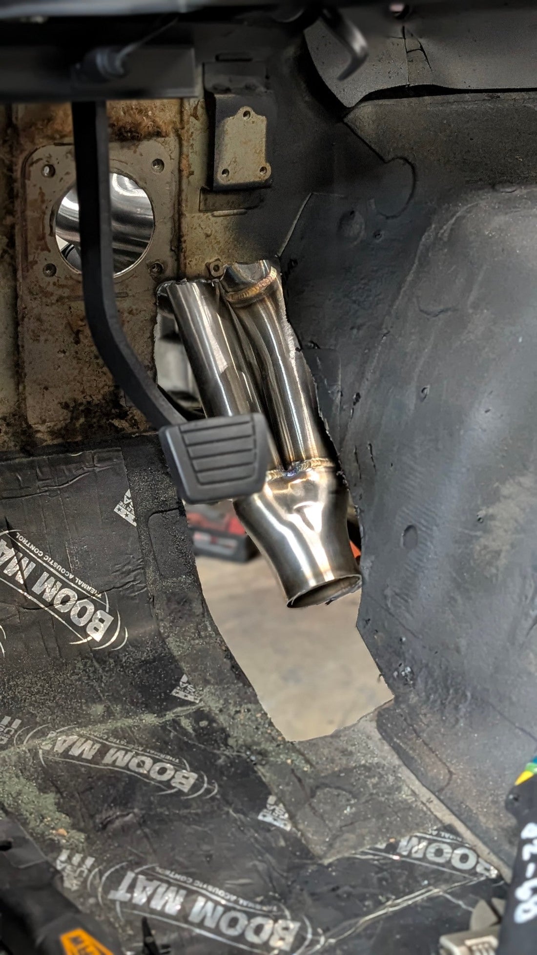 Header tubes and collector visible through an unfinished firewall and footwell opening beside a pedal.